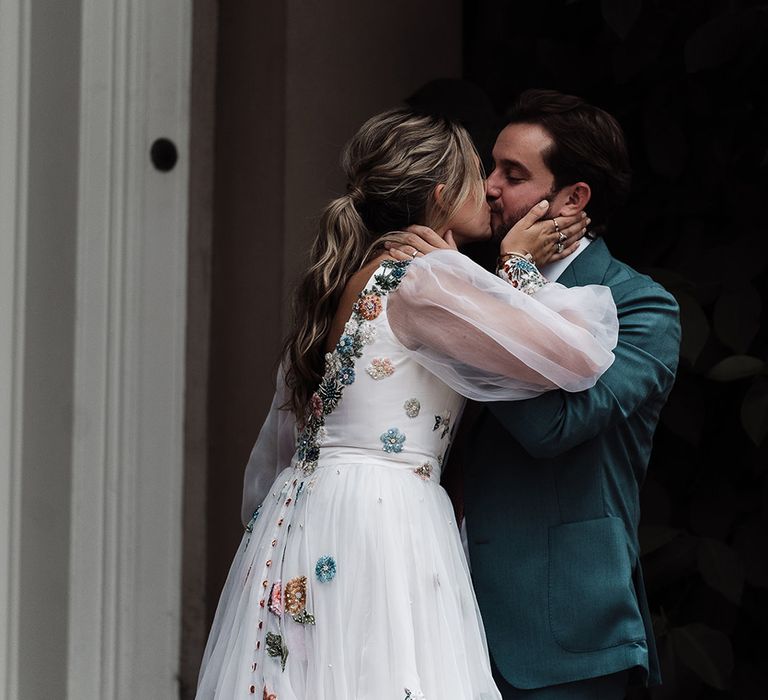 Bride in long mesh sleeve Victoria Sanders wedding dress with low back and floral embroidery kisses groom in teal suit in the grounds of Iscoyd Park