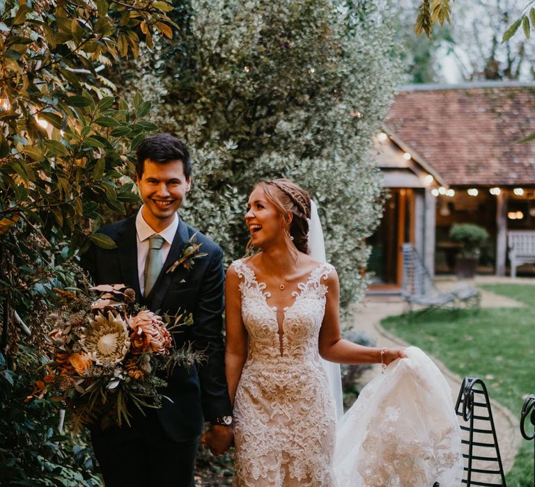 Bride in braided updo and lace wedding dress holding tulle train walks with groom in dark suit and sage green tie holding large bridal bouquet as they walk through gardens