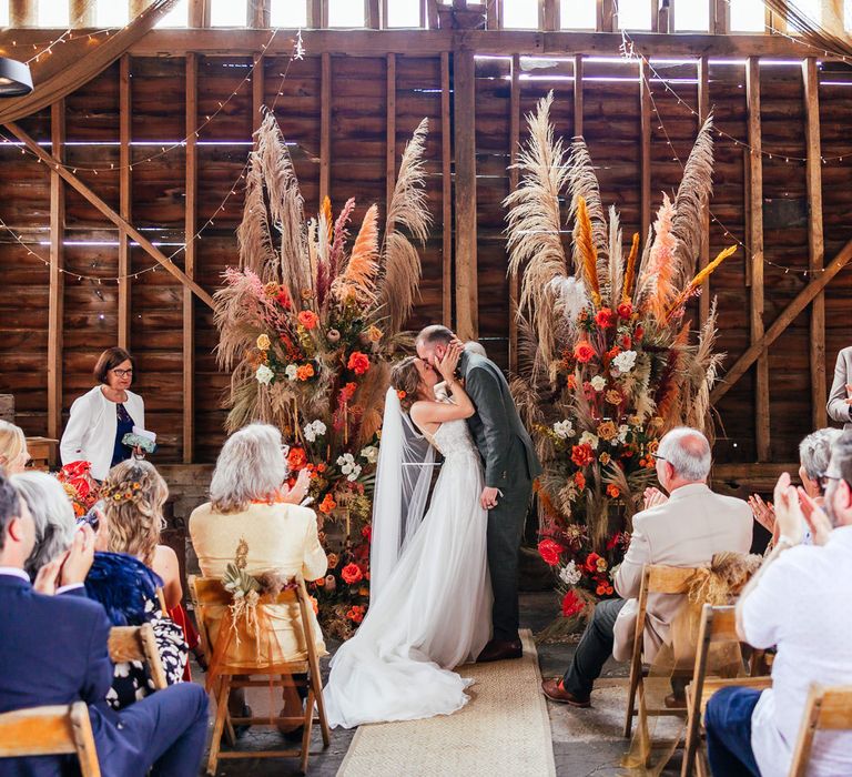 Bride in lace wedding dress with train kisses groom in green suit during festival style barn wedding ceremony with pampas grass and fairy lights as guests applaud 