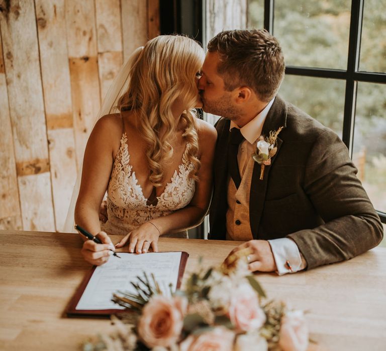 Bride in lace wedding dress and veil sits kissing groom in dark suit with white rose buttonhole as they sign marriage registry at barn wedding