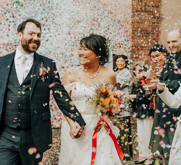 Groom in blue check three piece suit and light pink tie walks holding hands with bride in lace off the shoulder wedding dress and pearl necklace holding mixed bouquet tied with red ribbon as guests throw multi coloured confetti