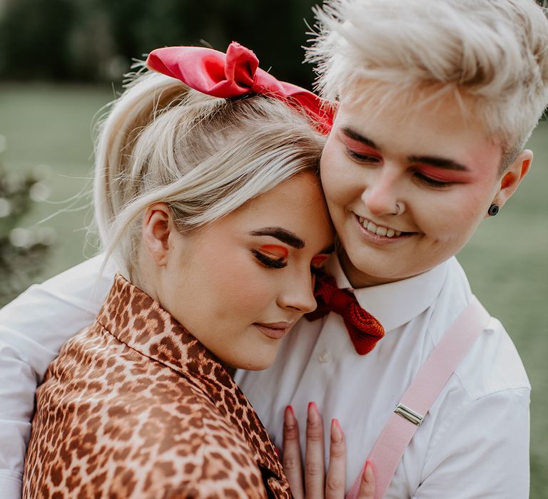 Two brides with red eye makeup embracing with pink braces, red ribbon hair accessory and leopard print jacket 