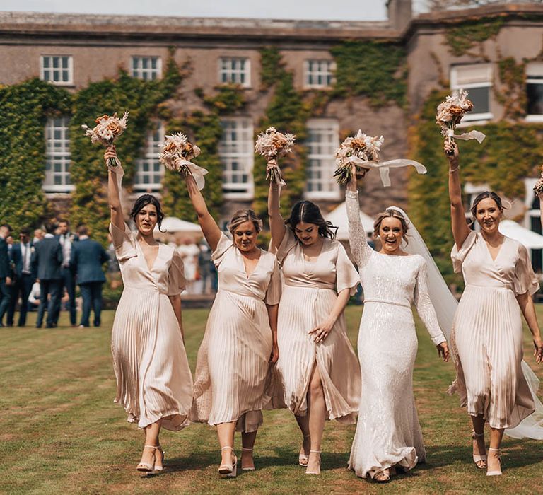 Bride in beaded wedding dress with long sleeves lifts her bouquet in the air as she walks with five bridesmaids in pale pink pleated dresses with butterfly sleeves