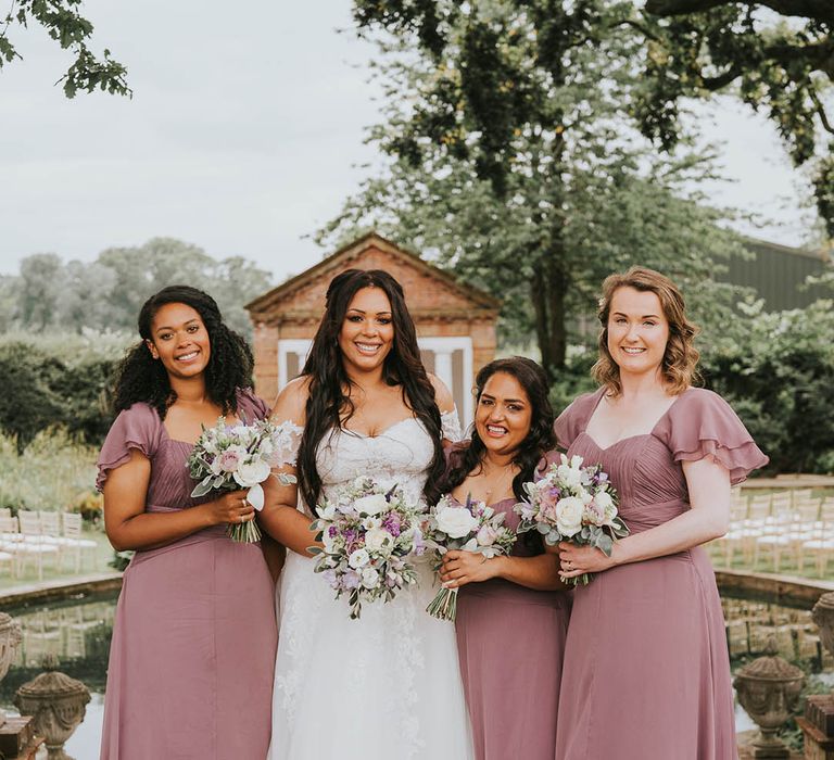 Bride and bridesmaids hold purple and white bouquets with bride in off the shoulder lace dress and bridesmaids in vintage pink dresses 