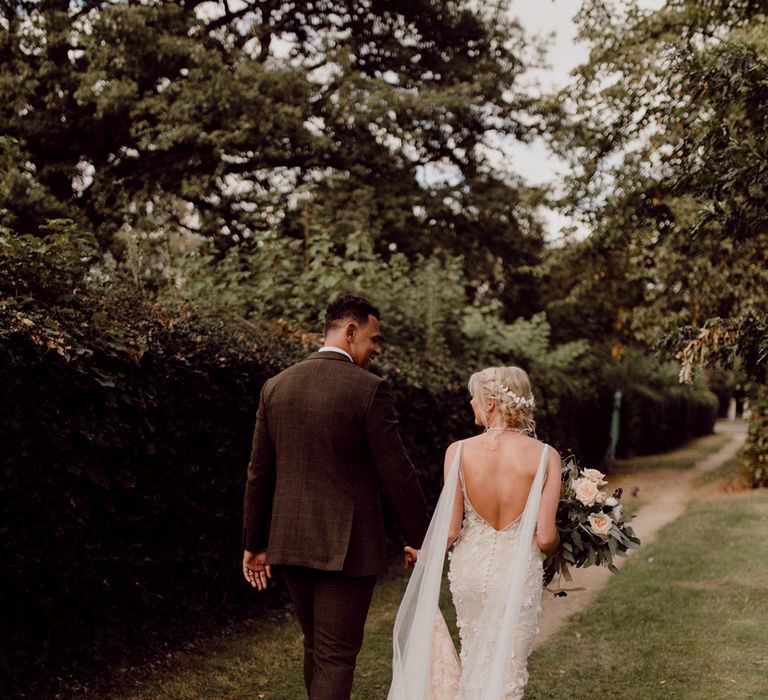 Bride with blonde braided updo and white flower hair accessory with Made With Love gown walks with groom in brown suit