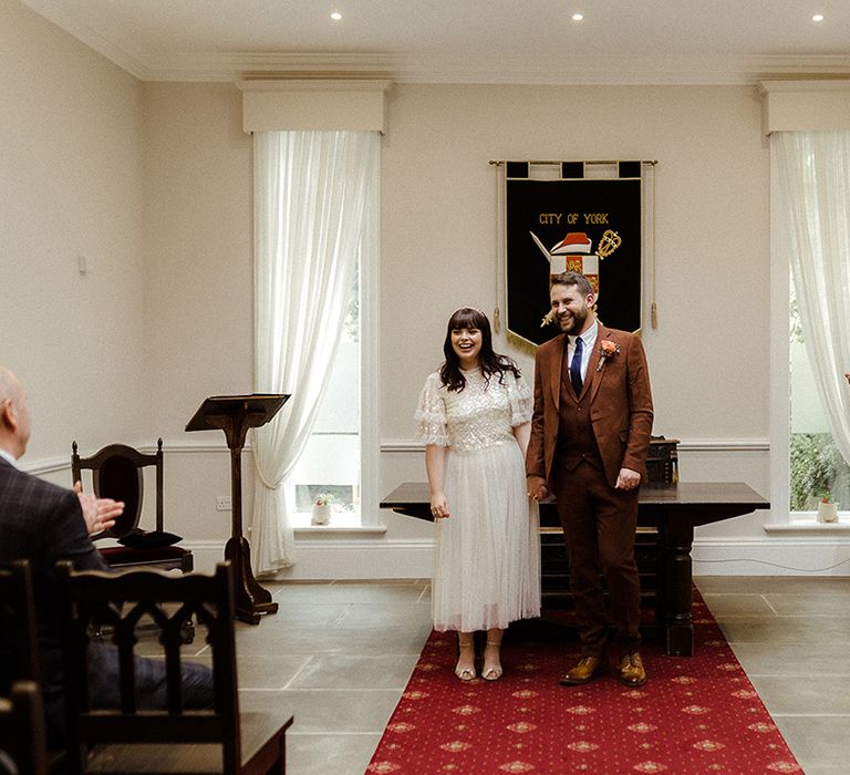 Bride in Needle and Thread bridal separates stands holding hands with the groom in brown suit after being announced husband and wife