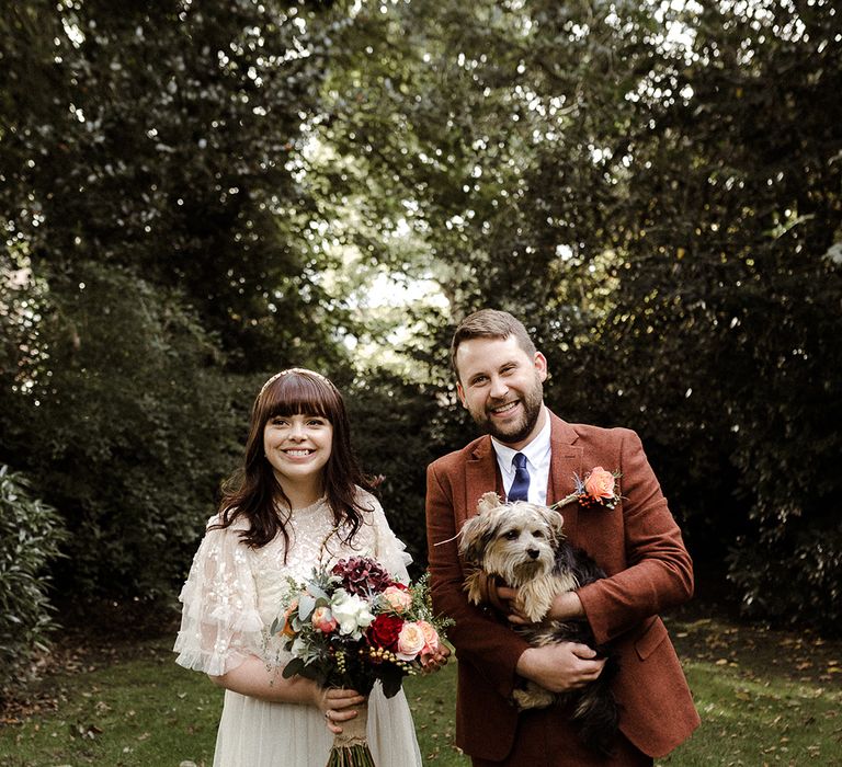 Bride and groom stand smiling together with the groom holding their pet dog