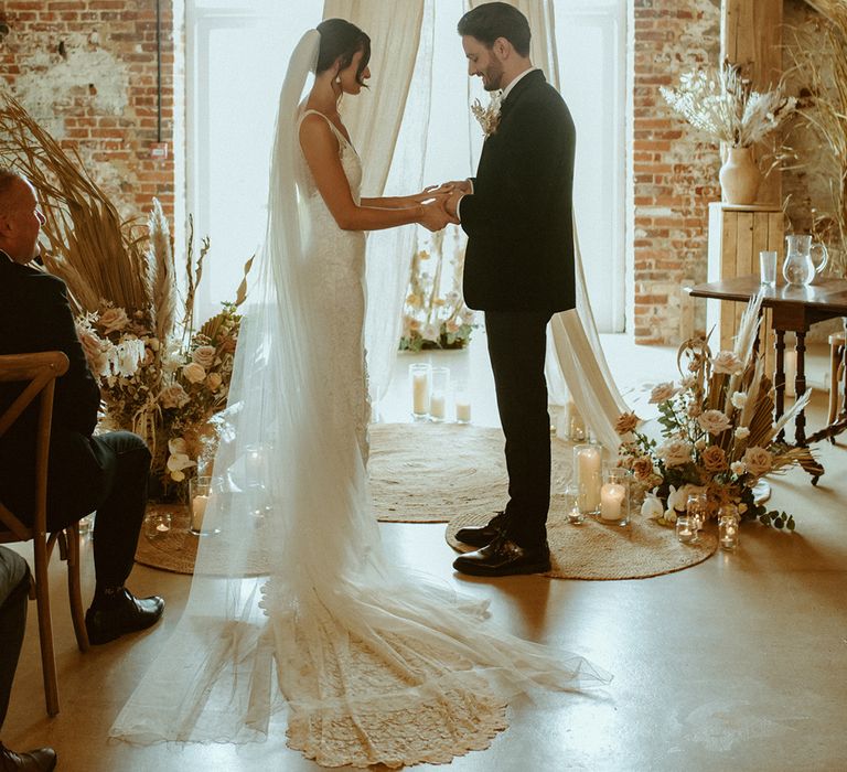 Bride and groom hold hands at the altar with white wedding drapes, dried flowers and candle for boho barn wedding