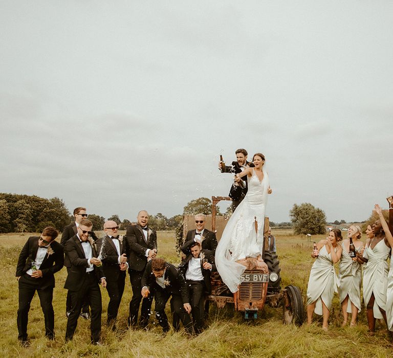 Bride and groom pop champagne on top of their tractor wedding transport with large wedding party drinking together 