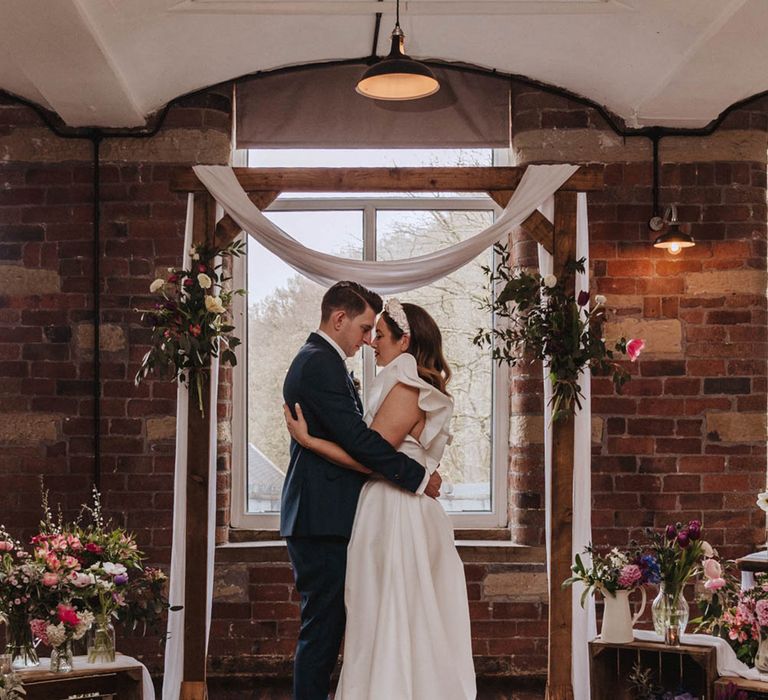 Bride and groom embrace at the altar decorated with crates of colourful spring flowers and wooden frame with white wedding drapery 