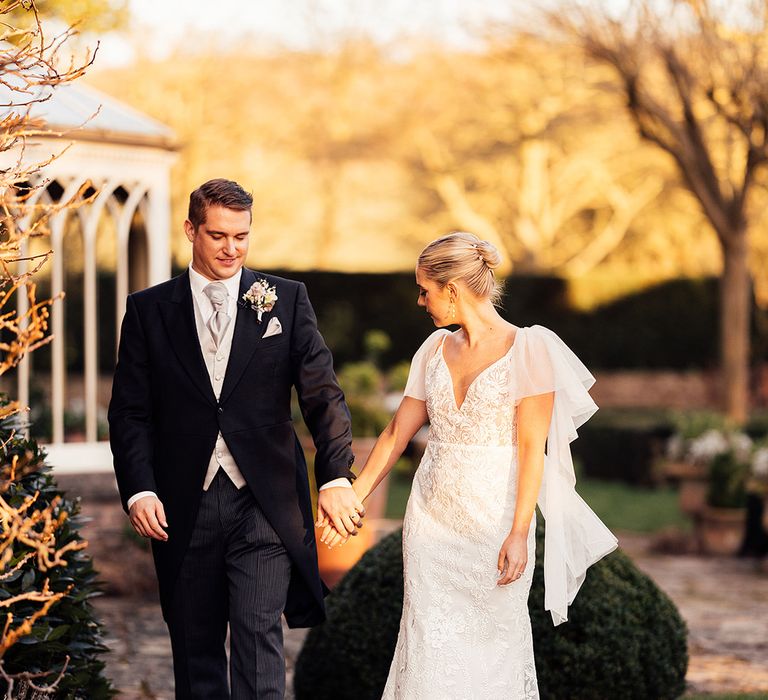 Bride in lace wedding dress with ruffle sleeves holding hands with groom in three piece morning suit with grey waistcoat and tie