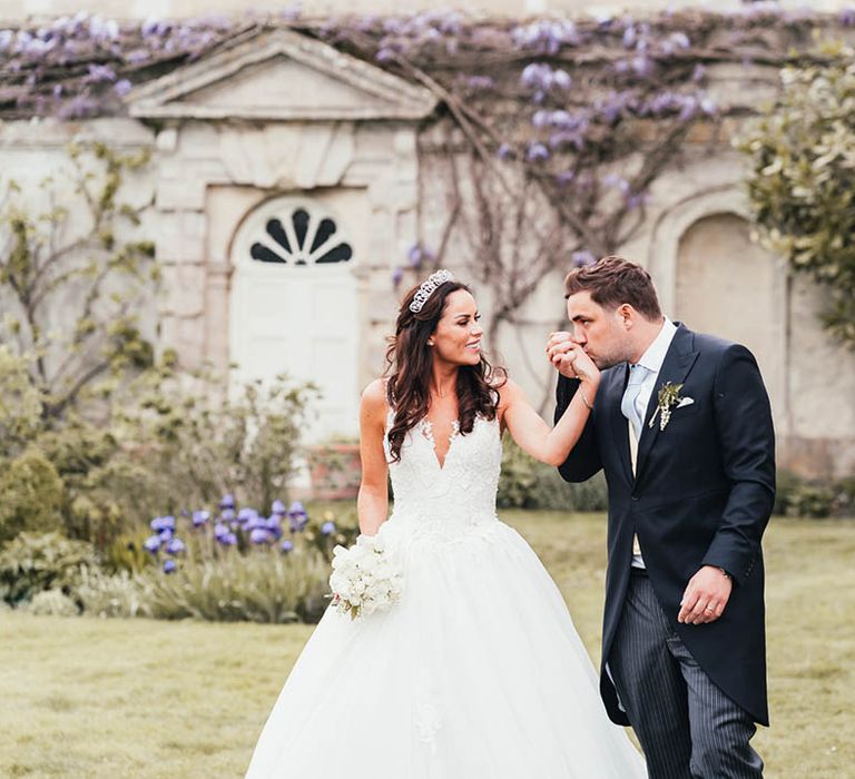 Groom in morning suit with pale blue tie kisses the hand of the bride as they walk together 