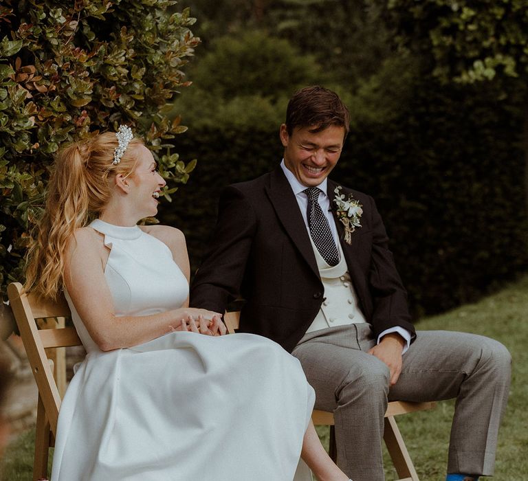 Bride in halter neck wedding dress with pearl embeliished wedding shoes and pearl headband laughing with groom in morning suit 