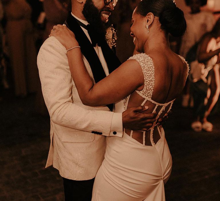 Bride in pearl detail open back wedding dress and groom in white tux doing their first dance