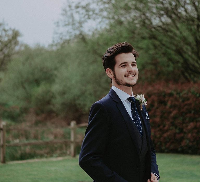 Groom in dark blue suit with polka dot blue and white tie smiles as he waits for the bride 