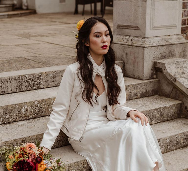 East Asian bride with long wavy hair wearing a flower in har hair, red lipstick, a satin slip wedding dress and a white leather jacket sitting on the steps at Gorse Hill