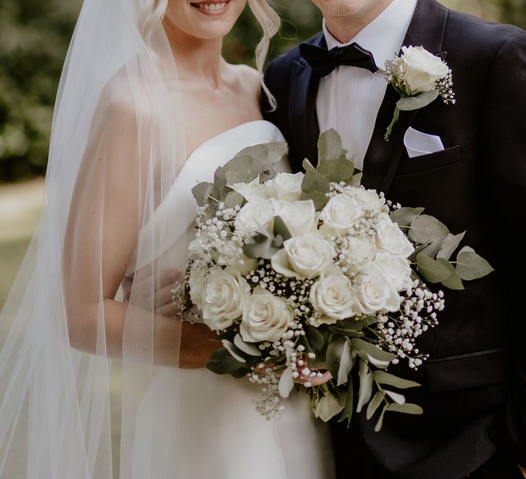 Bride and groom smile at the camera with the bride holding a white rose bouquet and the groom with a matching white rose buttonhole 