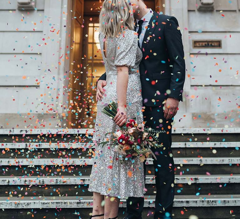 Bride & groom kiss beneath colourful confetti as bride wears silver mini wedding dress with short sleeves and back block heels 