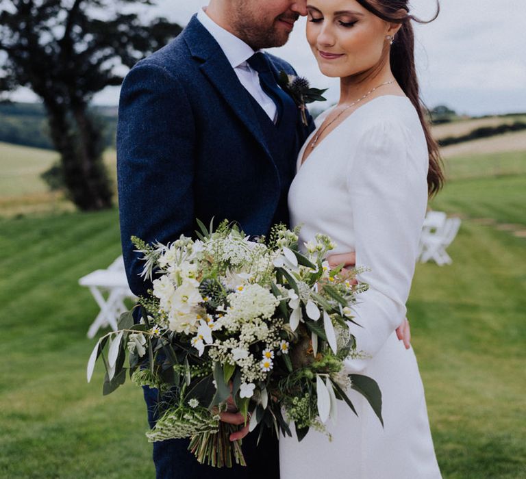 Groom in blue suit embraces the bride wearing a long sleeved wedding dress holding an all white flower bouquet 