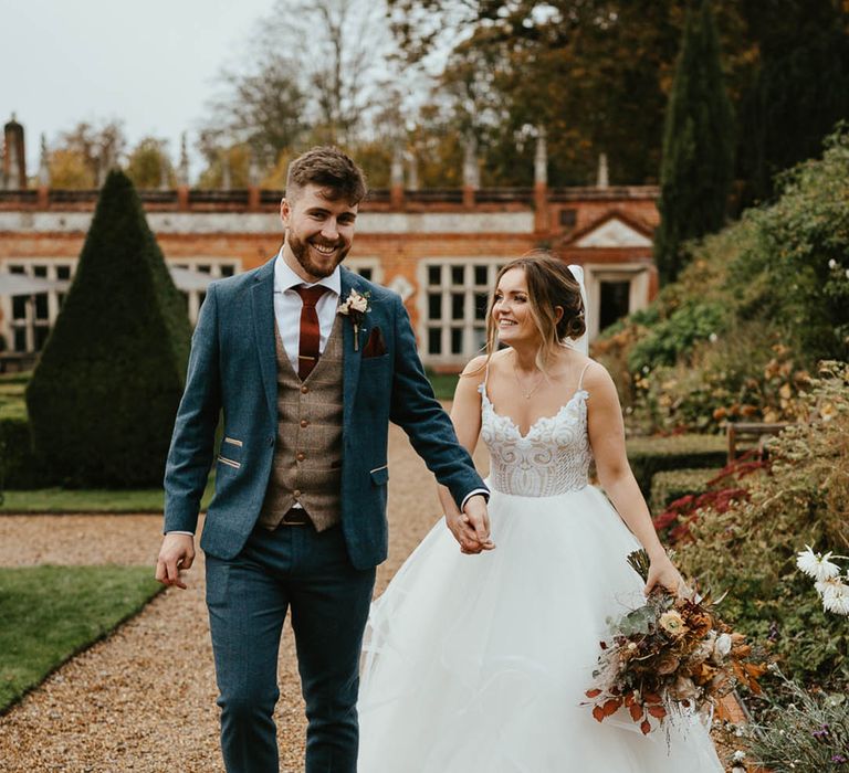 Groom in blue suit with brown waistcoat and burnt orange tie with bride in layered tulle wedding dress walking together around Oxnead Hall 