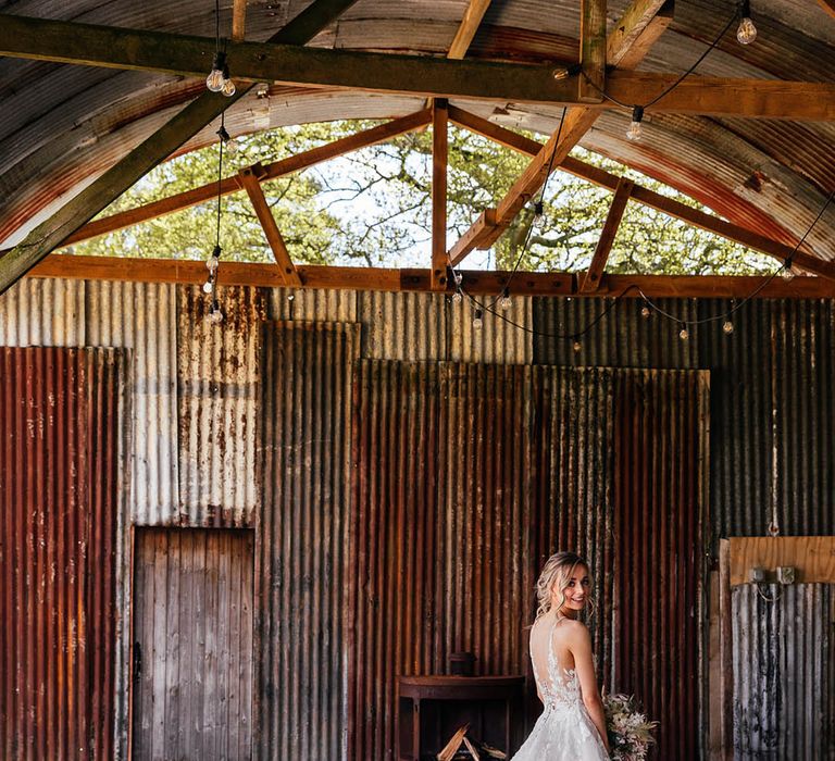 Bride in Madison James wedding dress with long train standing in barn wedding venue with festoon lighting 