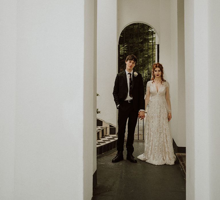 Bride wears lace wedding dress with long sleeves and V-shaped neckline as she holds her grooms hand in brown suit and white polkadot shirt