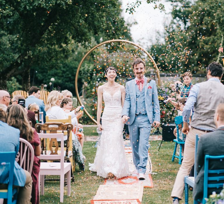 Groom wears three piece dusty blue suit with pink shirt and bow-tie as he walks with his bride through bright confetti 