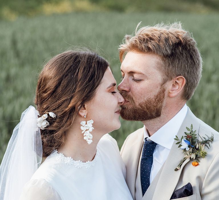 Groom in a three piece cream suit with a dark blue tie kisses the bride on the nose who wears a vintage wedding dress 