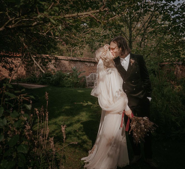 Bride holds dried floral bouquet and kisses her groom in a green suit during outdoor wedding 