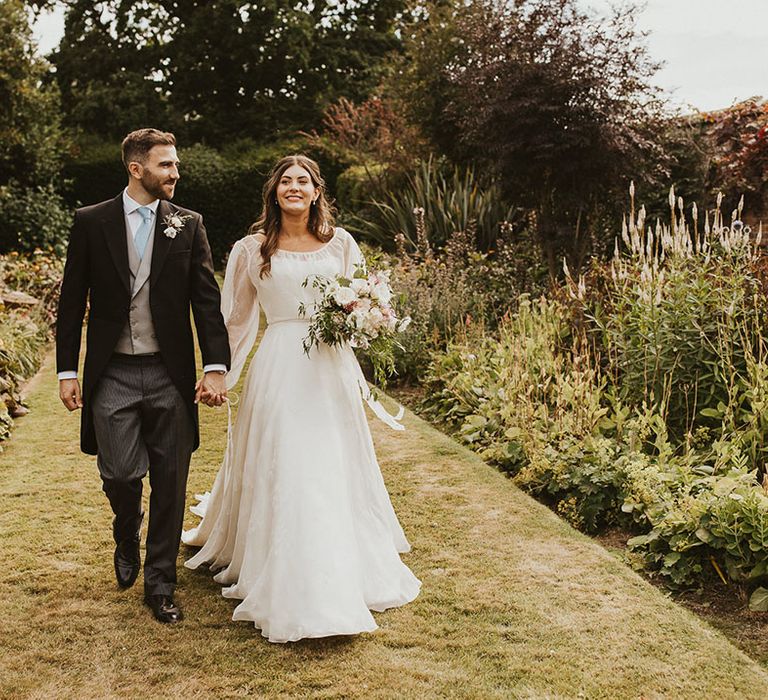 Groom in a morning suit with a pale blue tie and light grey waistcoat walking holding hands with the bride 