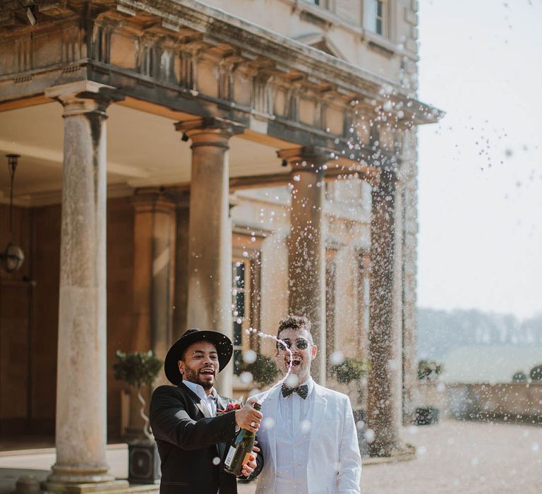 A groom in a black suit and hat with the groom in a white suit popping a bottle of no alcohol champagne to celebrate their wedding day 