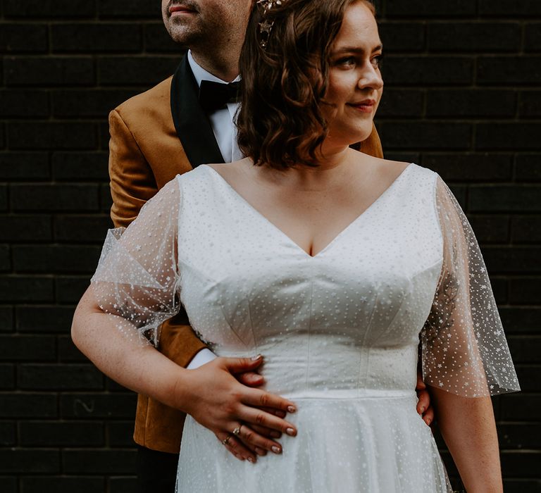 Bride and Groom pose in front of dark brick wall wearing a burnt orange velvet blazer and celestial wedding dress and crown