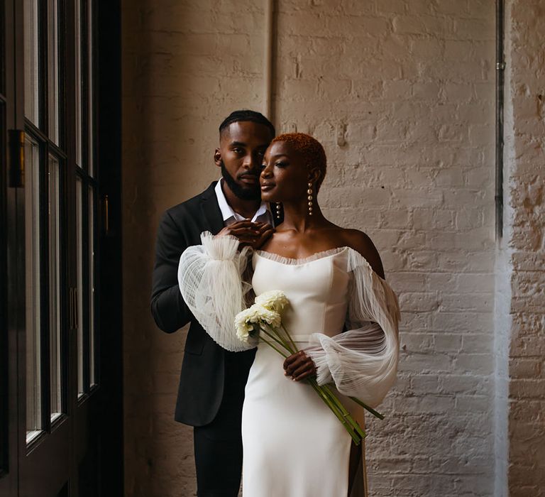 Groom in stylish skinny black tux and bride in off shoulder wedding dress with leg slit and puff lace sleeves holding a bunch of white dahlias posing at 100 Barrington