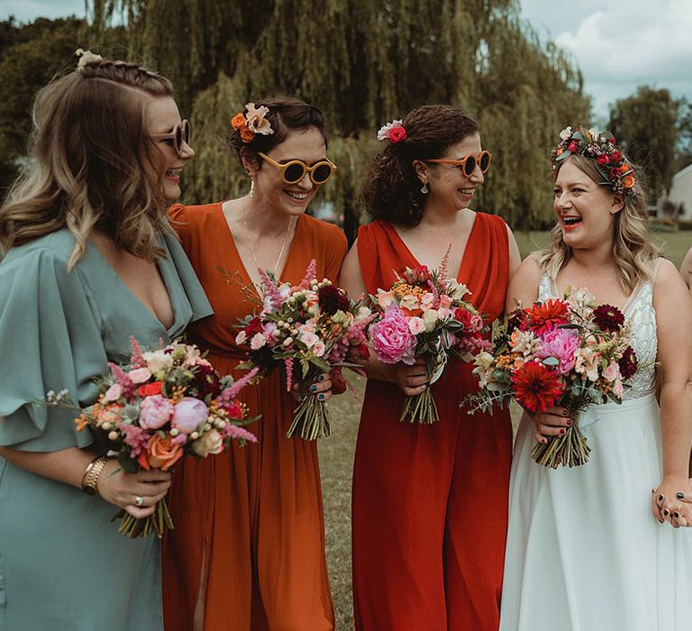 Bridesmaids in mismatched dresses and rounded sunglasses with colourful bouquets smiling with the bride 