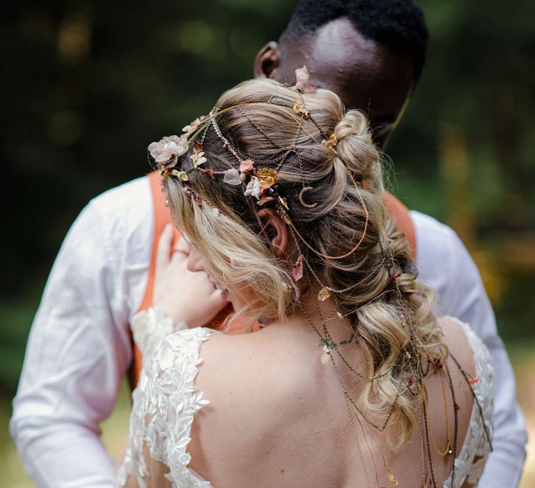 Bride in open back detail wedding dress with lace floral detailing and headpiece with delicate details and mix of soft gold & bronze metals and groom embracing her with orange waistcoat and white shirt