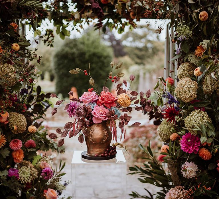 Luxe autumnal flower arch surrounding single tier iced wedding cake with intricate leaf details in the style of an aged golden vase filled with handmade wafer paper florals, pink roses, red roses and yellow carnations 