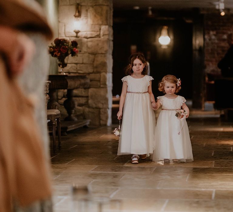 Flower girls in white floor length dresses with embellished belts walk down the aisle 