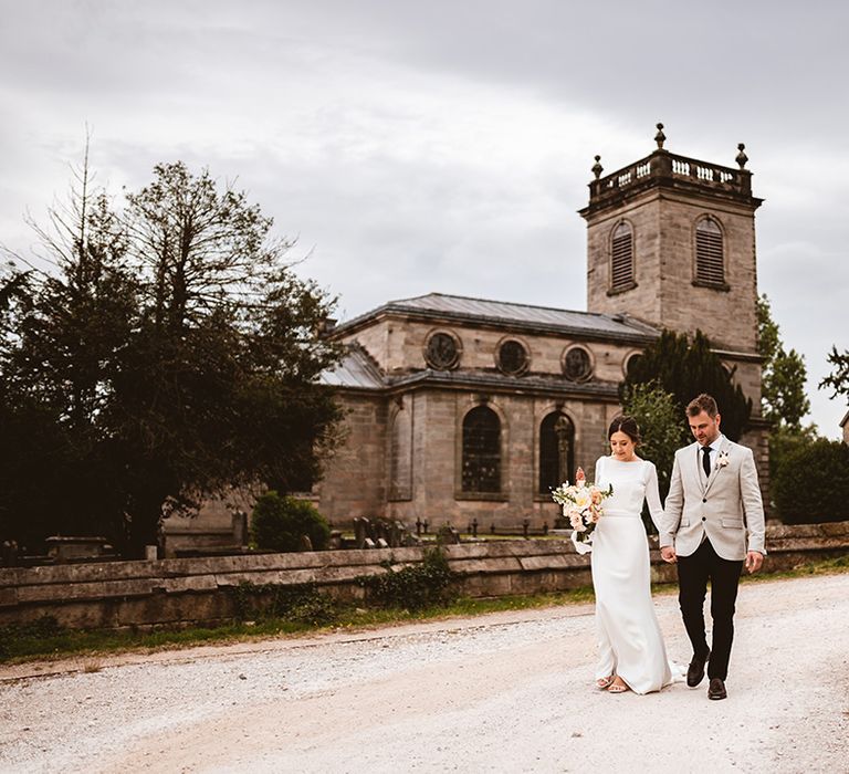 Groom in black suit trousers, grey blazer and light pink rose boutonniere and bride in long satin wedding dress with bridal cover up semi sheer crop top walking around The Orange Ingestre wedding venue