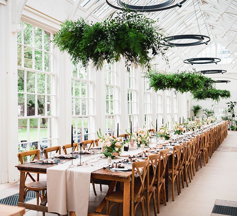 Large glasshouse wedding venue reception room with long wedding tablescape, with neutral off-white table runners, rose and foliage floral arrangements, pink and black tapered candles, black candlestick holders and suspended foliage decorations