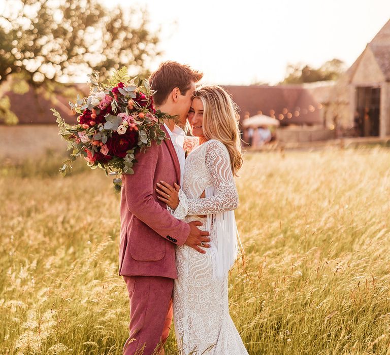 Bride in long sleeve lace boho wedding dress with tassels and groom in pink corduroy suit with pink and red rose, foliage and dried flower boutonniere embracing at Old Gore Barn