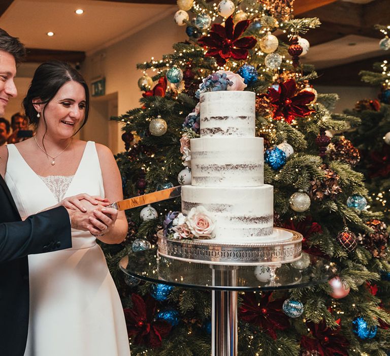 Three tier chocolate cake semi-naked frosted wedding cake being cut by the bride and groom in front of the Christmas tree  