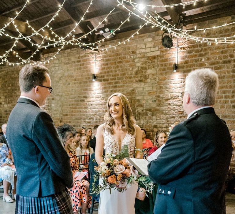 Bride holds orange bridal bouquet and looks lovingly toward her groom during Christian wedding ceremony 