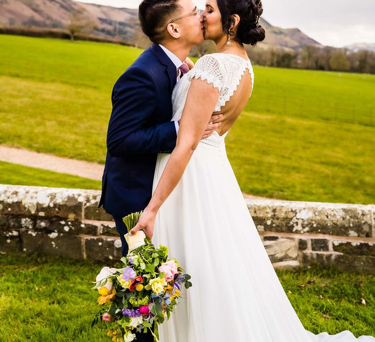 Bride wears lace open back wedding dress whilst holding colourful floral bouquet and kissing her groom outdoors 