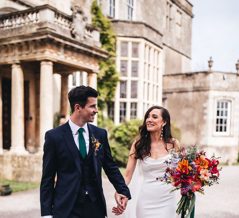 Bride in fitted cowl neck satin Kyha wedding dress with pearl earrings holding colourful bouquet walking with groom in navy suit outside Elmore Court