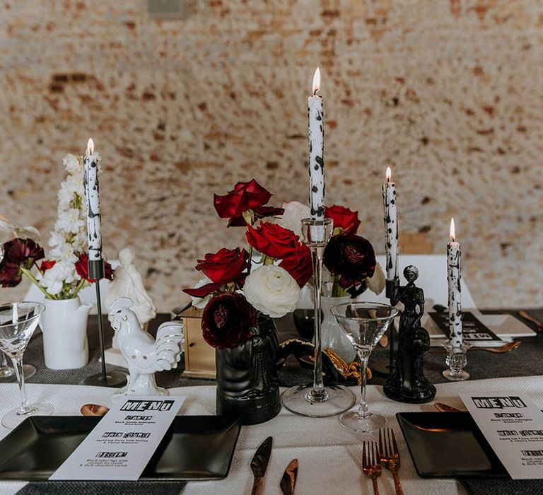 Contemporary, edgy wedding tablescape with black red and white wedding decor - black and white tapered candles in clear candlestick holders, red roses, red carnations and white carnations, small black and white sculptures, and gold cutlery 