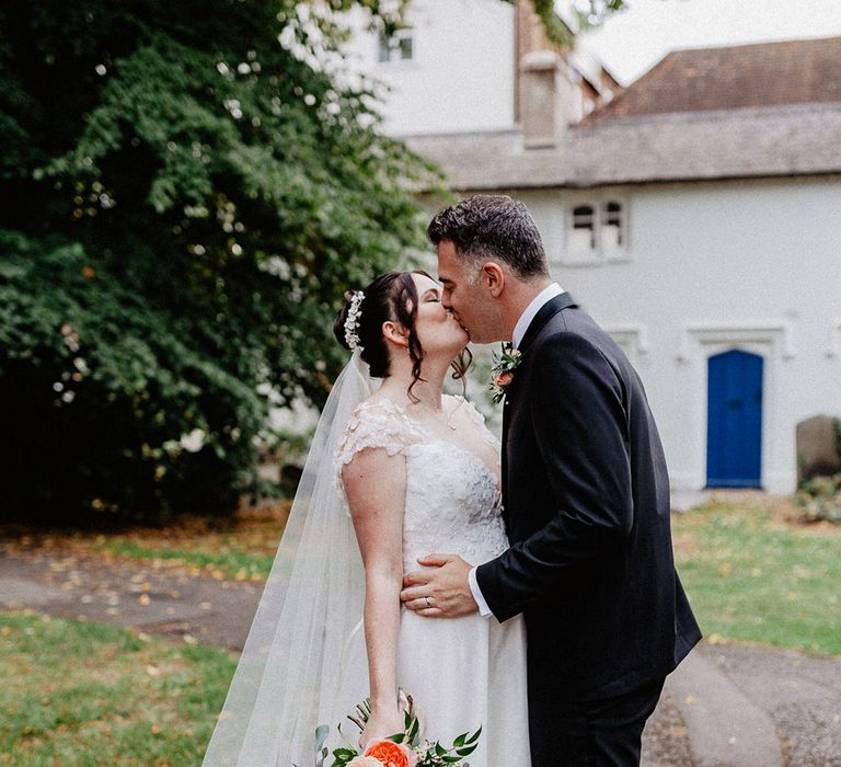 The bride with a flower hairpiece and flower wedding dress holding pastel wedding bouquet kisses the groom in black tie 