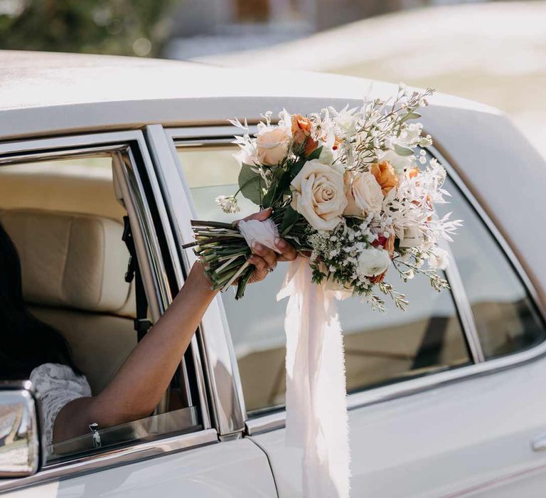 Bride holding bouquet with neutral toned garden roses, carnations, baby's-breath, eucalyptus and dried flowers out the window of classic white wedding car 