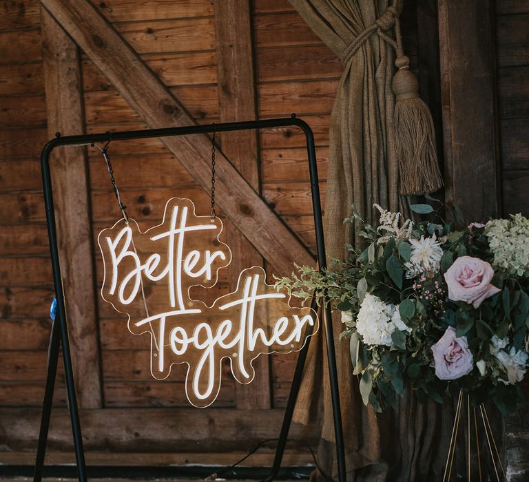 White neon sign reading 'Better Together' hanging on a black metal frame next to a pastel flower arrangement for wedding decor 