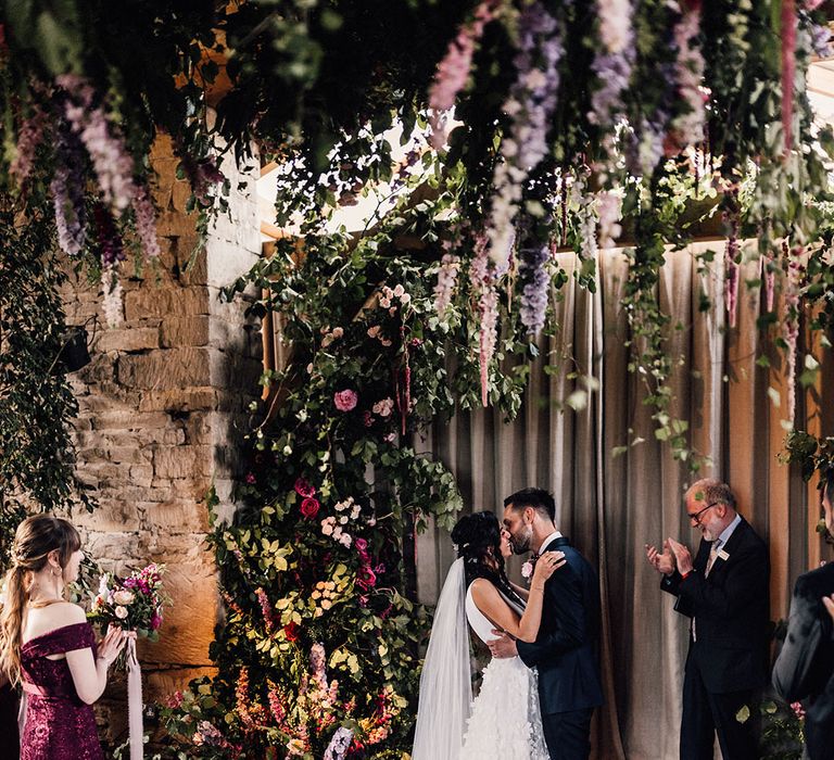 The bride and groom share their first kiss as a married couple surrounded by beautiful flowers at their barn wedding 