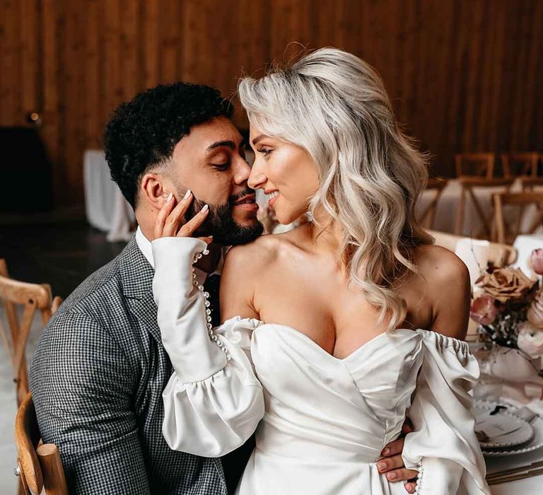 Bride in Nori Ivy off the shoulder long sleeve wedding dress with slight cowl neck and front leg slit sitting on groom's lap at rustic luxe wedding at Lodge Farm